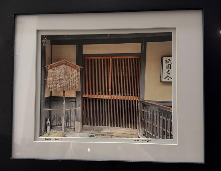 Kyoto Threshold by Carol Spungen Framed photograph of a traditional Japanese wooden storefront in Kyoto, featuring a sliding slatted door, weathered wooden signboard with Japanese writing, and muted beige walls.