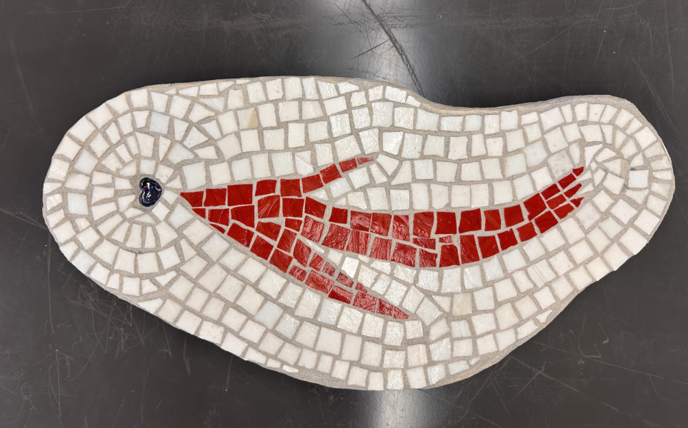 Oval-shaped mosaic artwork composed of small white tiles arranged in swirling patterns, forming a wave-like background. At the center, bright red tiles create the silhouette of a stylized fish swimming across the piece, with a small dark blue accent near the head. The mosaic rests on a dark surface, highlighting the contrast between the white and red tiles.