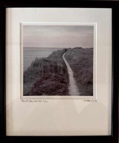 Boy with Stick, More Mesa by Bill Dewey Framed black-and-white photograph of a narrow dirt path winding along a coastal bluff above the ocean, with a small figure walking in the distance under an overcast sky.