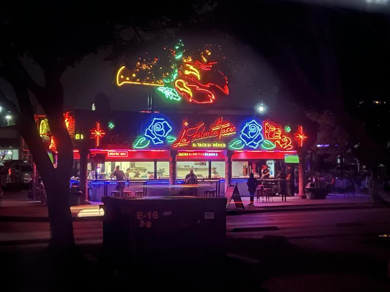 Tijuana Taqueria by Helene Schneider Neon-lit taco stand at night with colorful signs of peppers, roses, and flames, bright red and blue counter lighting, and people ordering food under glowing signage